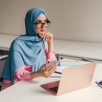 young pretty modern muslim woman in hijab working on laptop in office room, education online, remote work freelancer, using smatphone thinking